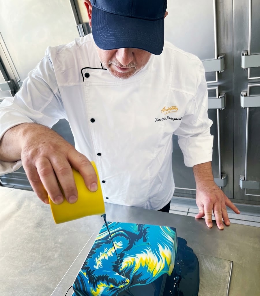 Demetrios pouring blue and yellow mirror glaze over a cake